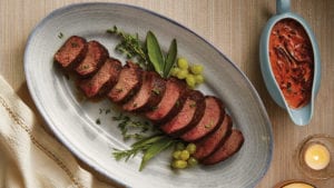 sliced venison loin on oval dish with thyme and green grapes and mushroom cassis sauce in ceramic bowl