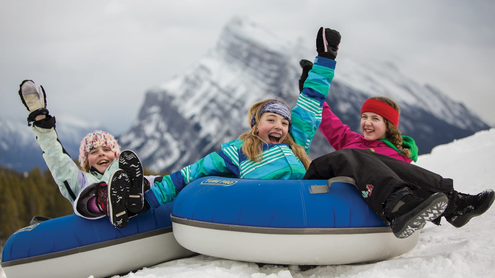 three young girls waving on snow tubes with mountains in background
