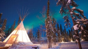 White tipi in snow-covered forest outside Yellowknife with northern lights and stars in sky