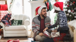 smiling bearded man in santa hat receiving gift from woman beside Christmas tree