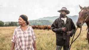 Cheryl Foggo standing in prairie field beside actor in cowboy costume with cattle in background