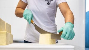 man wearing green vinyl gloves cutting block of white cheese with two-handed knife