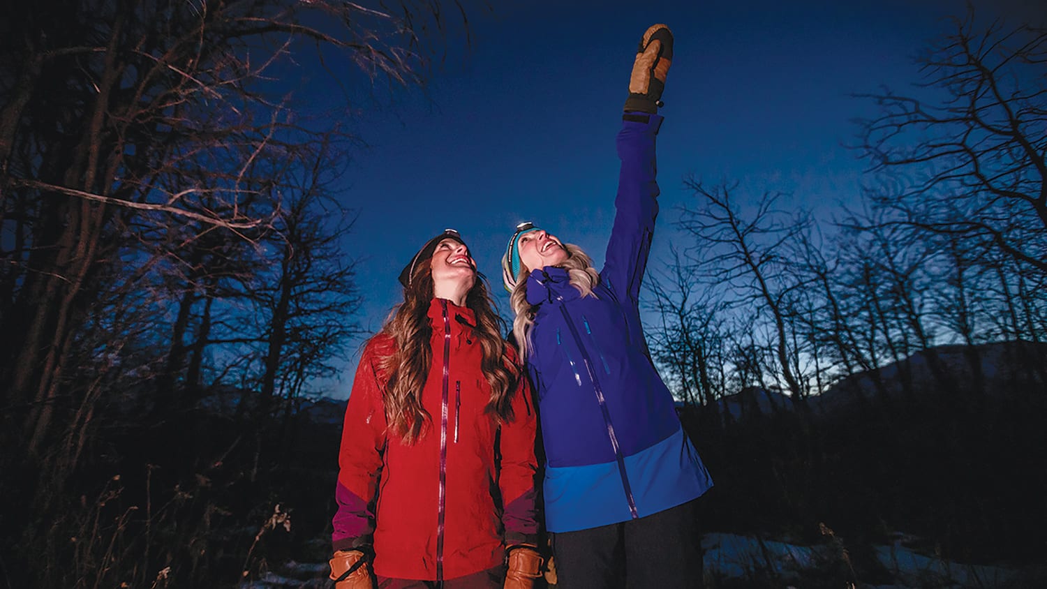Two women with winter clothes and headlamps pointing at night sky in Waterton Lakes National Park