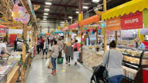 Customers walking through Alberta farmers market indoors lined by various vendor booths and stands
