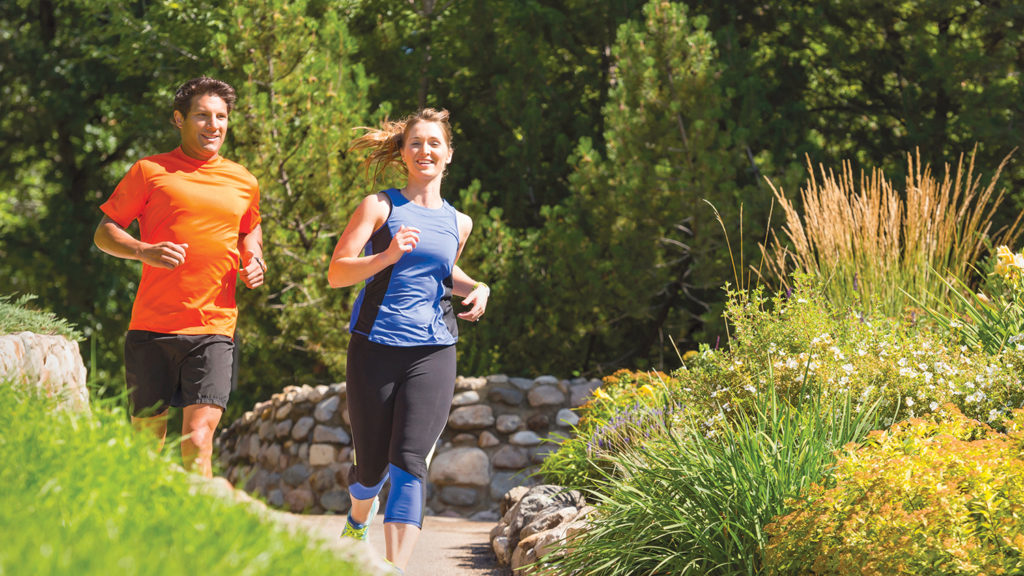 man and woman jogging on paved trail past lush greenery during visit to Saskatoon