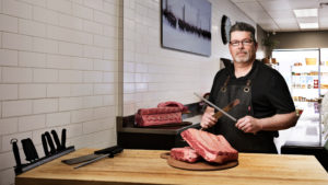 bearded man holding knives at Alberta butcher shop counter preparing to cut into meat