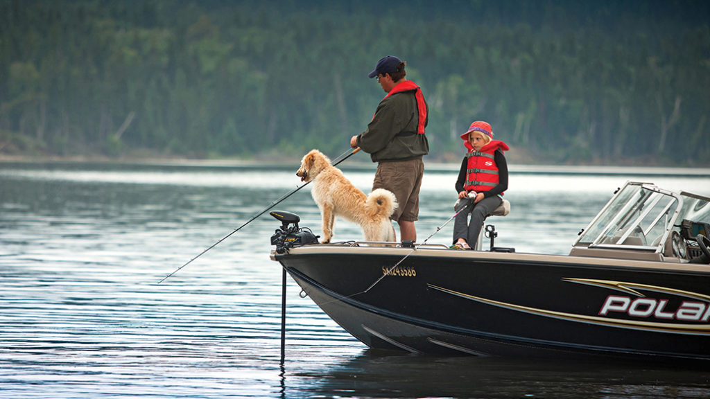 Man, boy and dog fishing from front of boat with forest in background