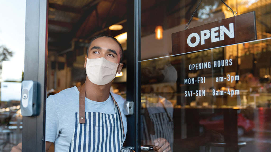 Man wearing mask and apron looking out door of cafe with opening hours written on it