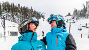 Man and younger girl smiling at one another wearing ski equipment with ski hill in background