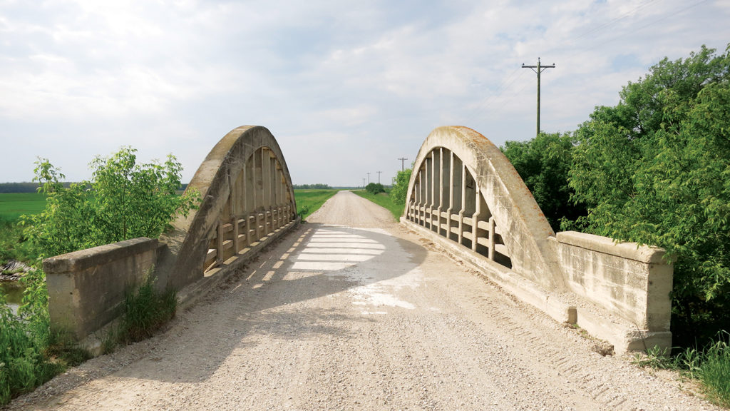 gravel road crossing a concrete arch bridge surrounded by scrub bushes and fields