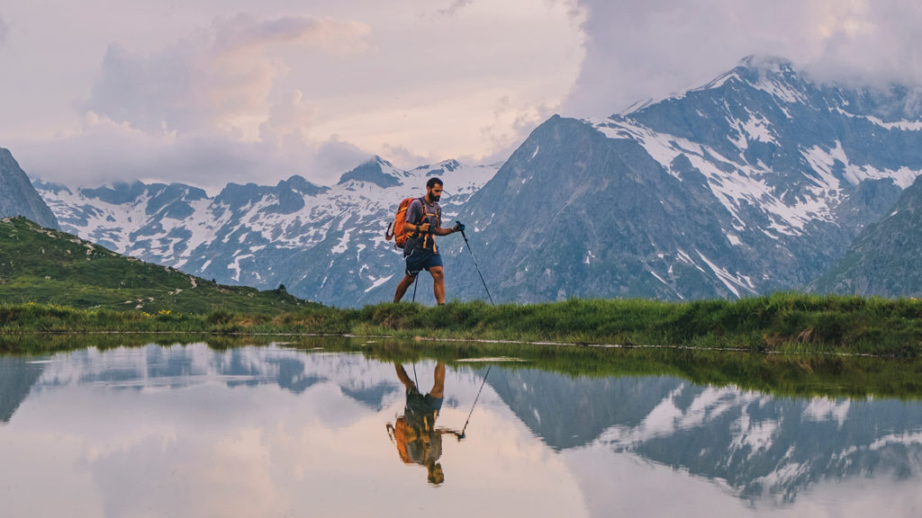 Man walking with backpack along shore of lake with alpine mountains in background