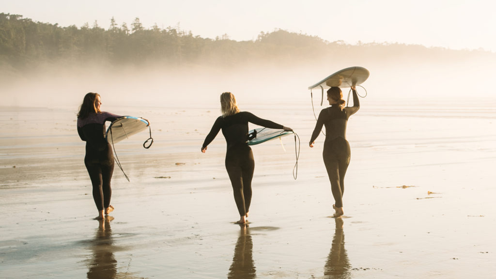 Three women carrying surf boards on foggy beach in Tofino